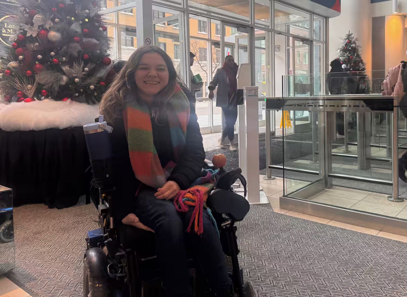 A young woman with a colorful scarf sits in a wheelchair in front of a decorated Christmas tree, near a glass entrance to a building.