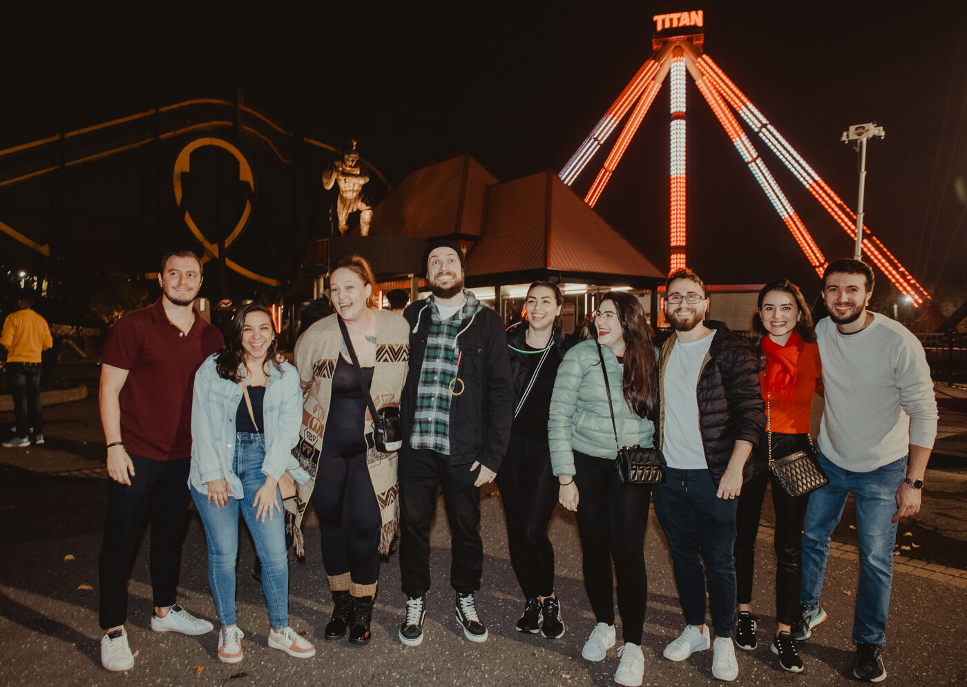 Nine friends pose with joy at a night-time amusement park, with colorful ride lights illuminating the background.