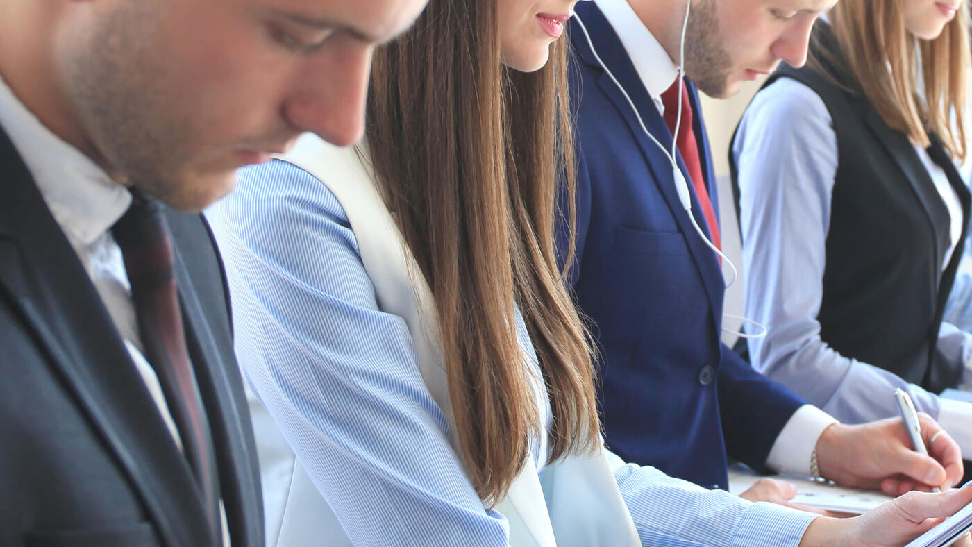 Two men and two women, all dressed in business formal attire, are engrossed in work on their digital devices at a meeting.