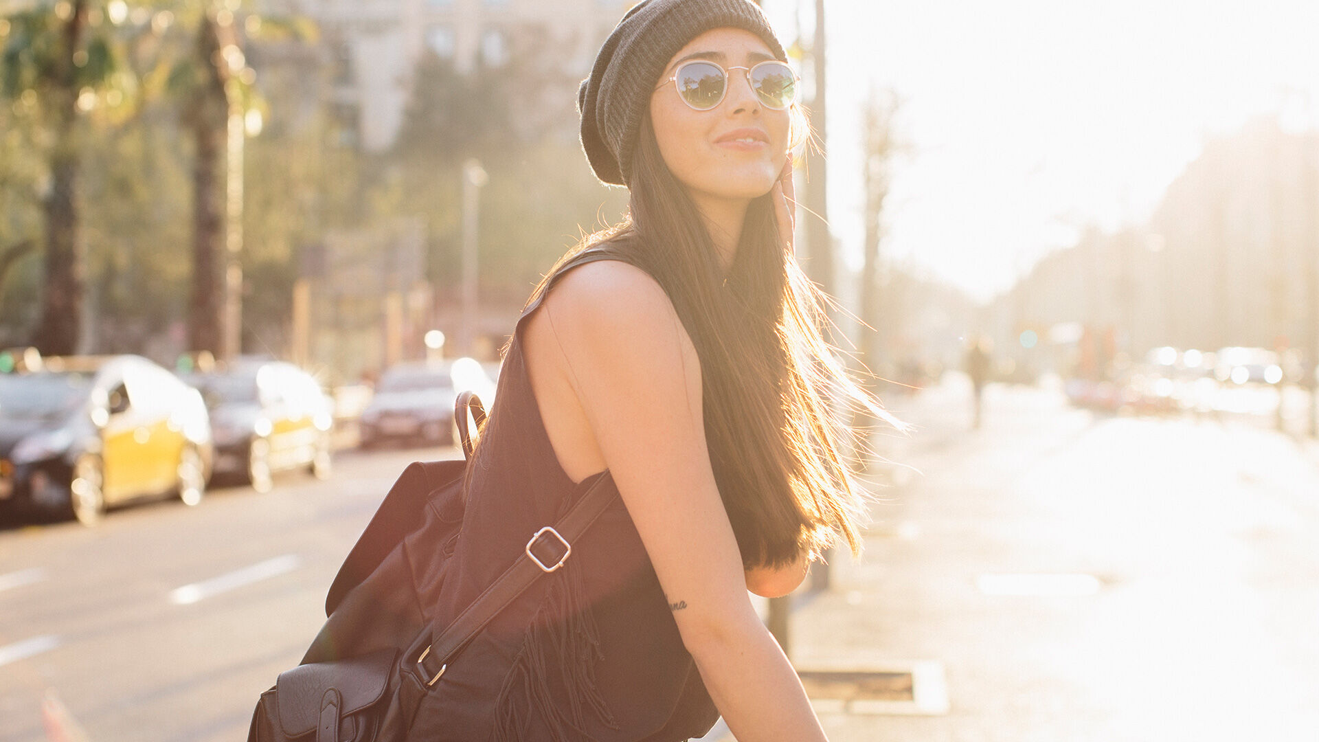Une femme élégante avec des lunettes de soleil et un bonnet marche dans une rue ensoleillée de la ville, dégageant une ambiance moderne et détendue.