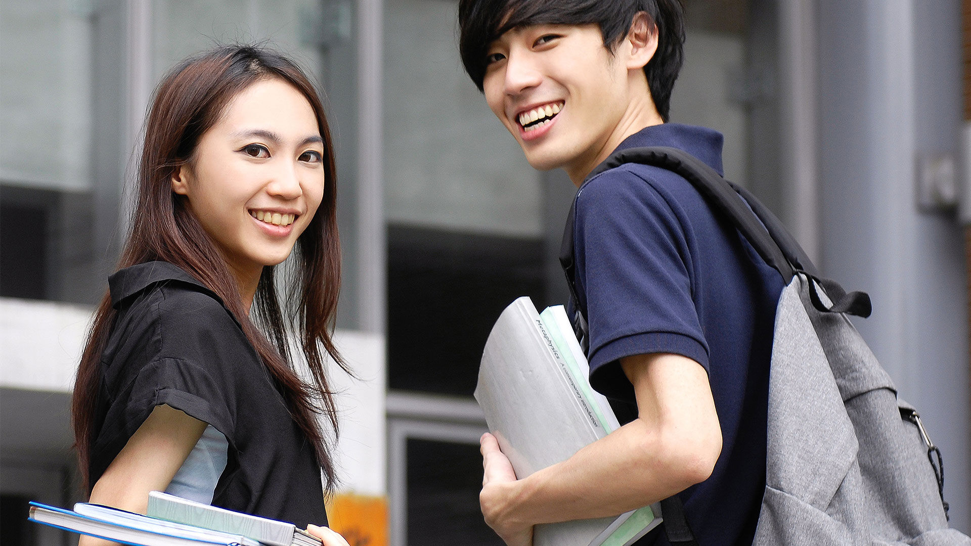 Deux étudiants joyeux avec des sacs à dos et des livres de cours marchent à l'extérieur d'un bâtiment universitaire.