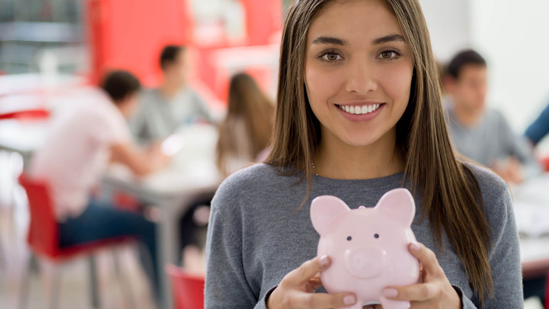 A smiling young woman in a grey sweater holds a pink piggy bank confidently in an office setting, suggesting financial planning or savings.