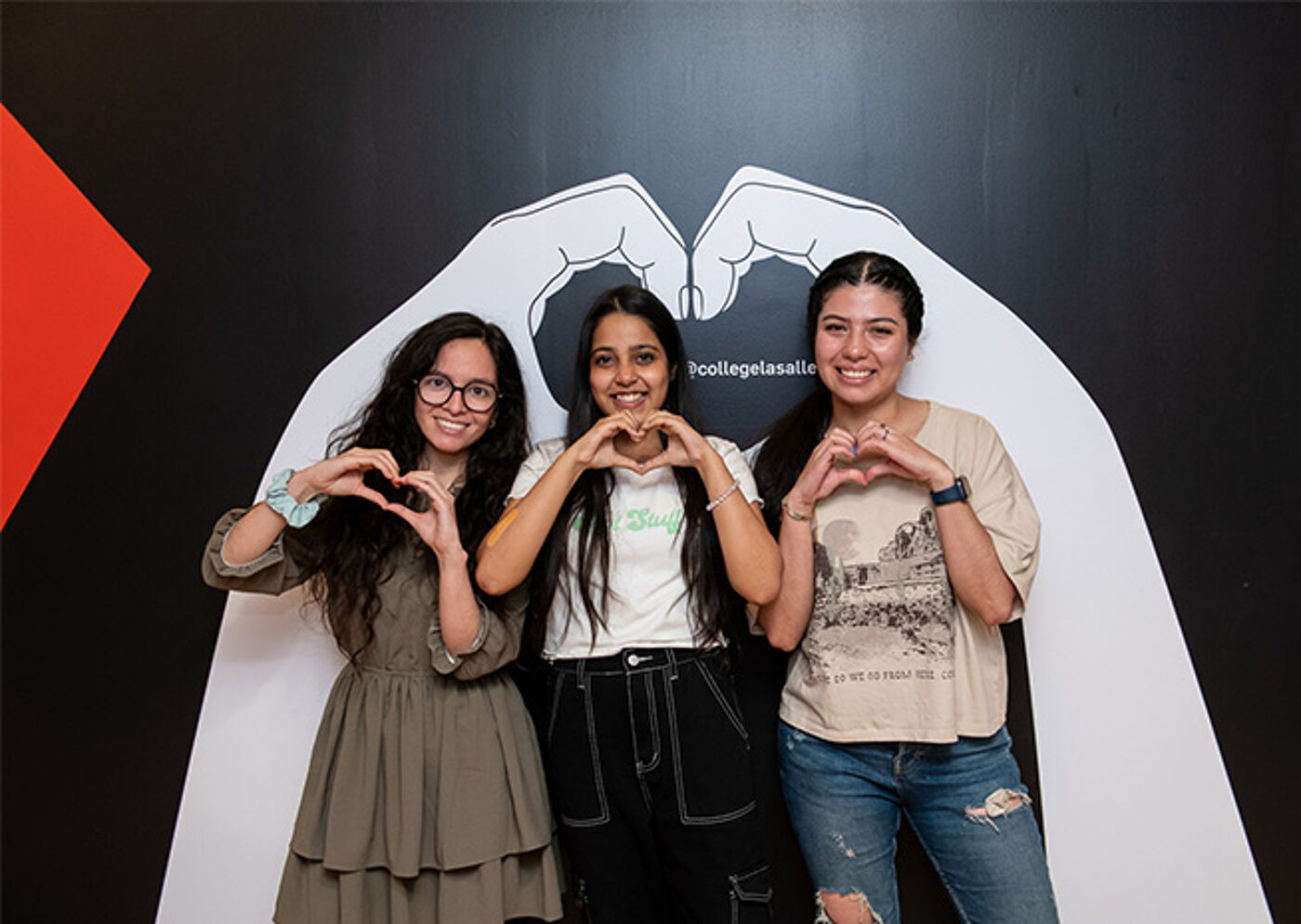 Trio of smiling women posing with hands in heart gestures, standing before a backdrop with wing illustrations.