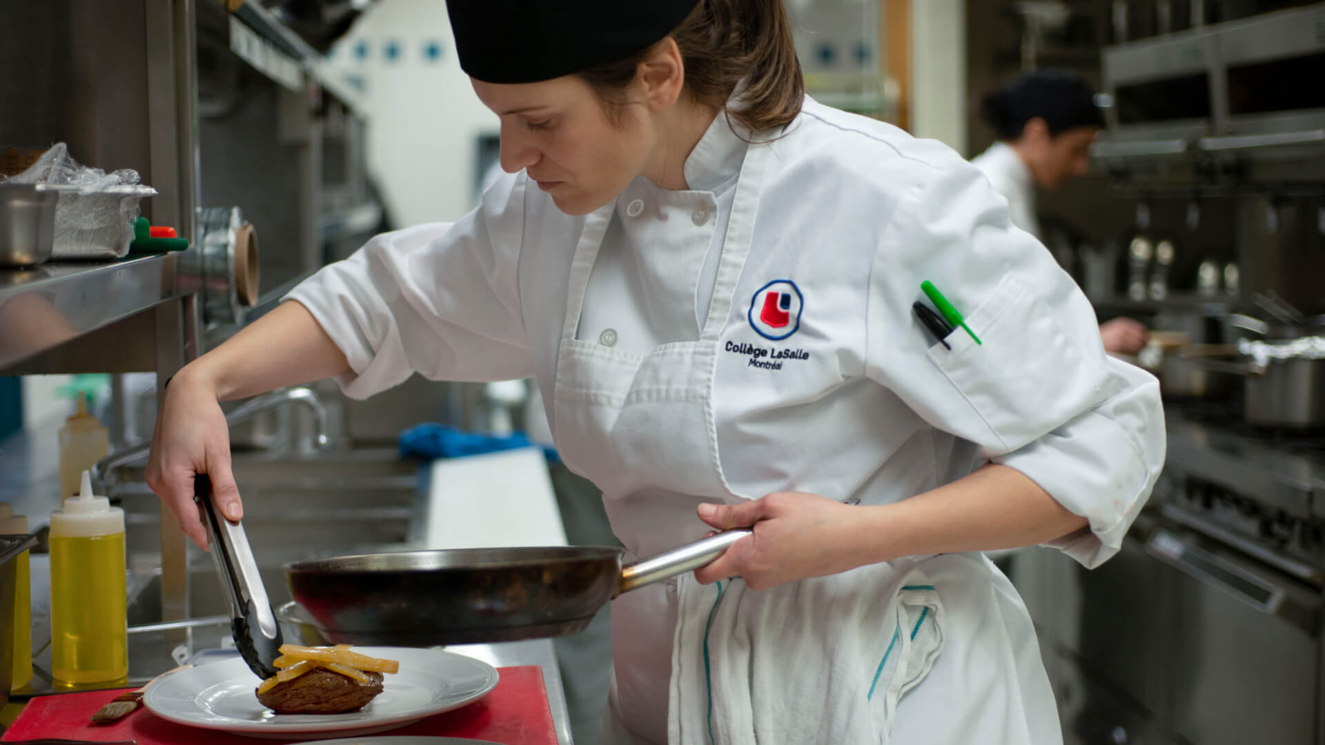 Une étudiante en cuisine dévouée au Collège LaSalle dresse habilement un plat, concentrée sur le détail dans une cuisine animée.
