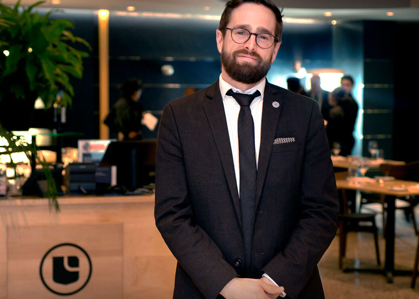 The image features a man wearing glasses, a suit, and a tie, standing in front of a bar or a restaurant. He is posing for a picture, possibly for a business or professional purpose.