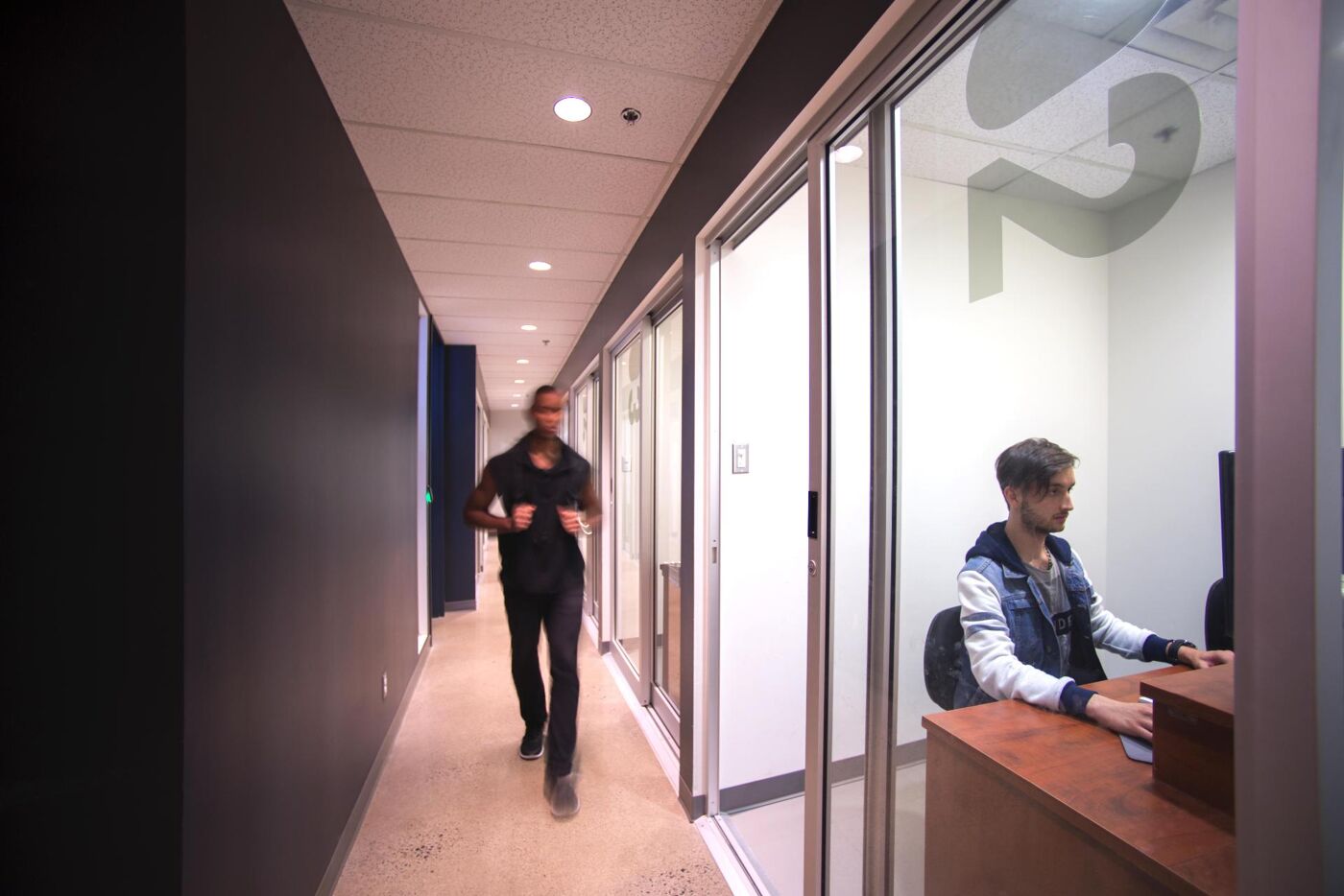  A man walks briskly through an office corridor while another sits at a workstation with a computer.