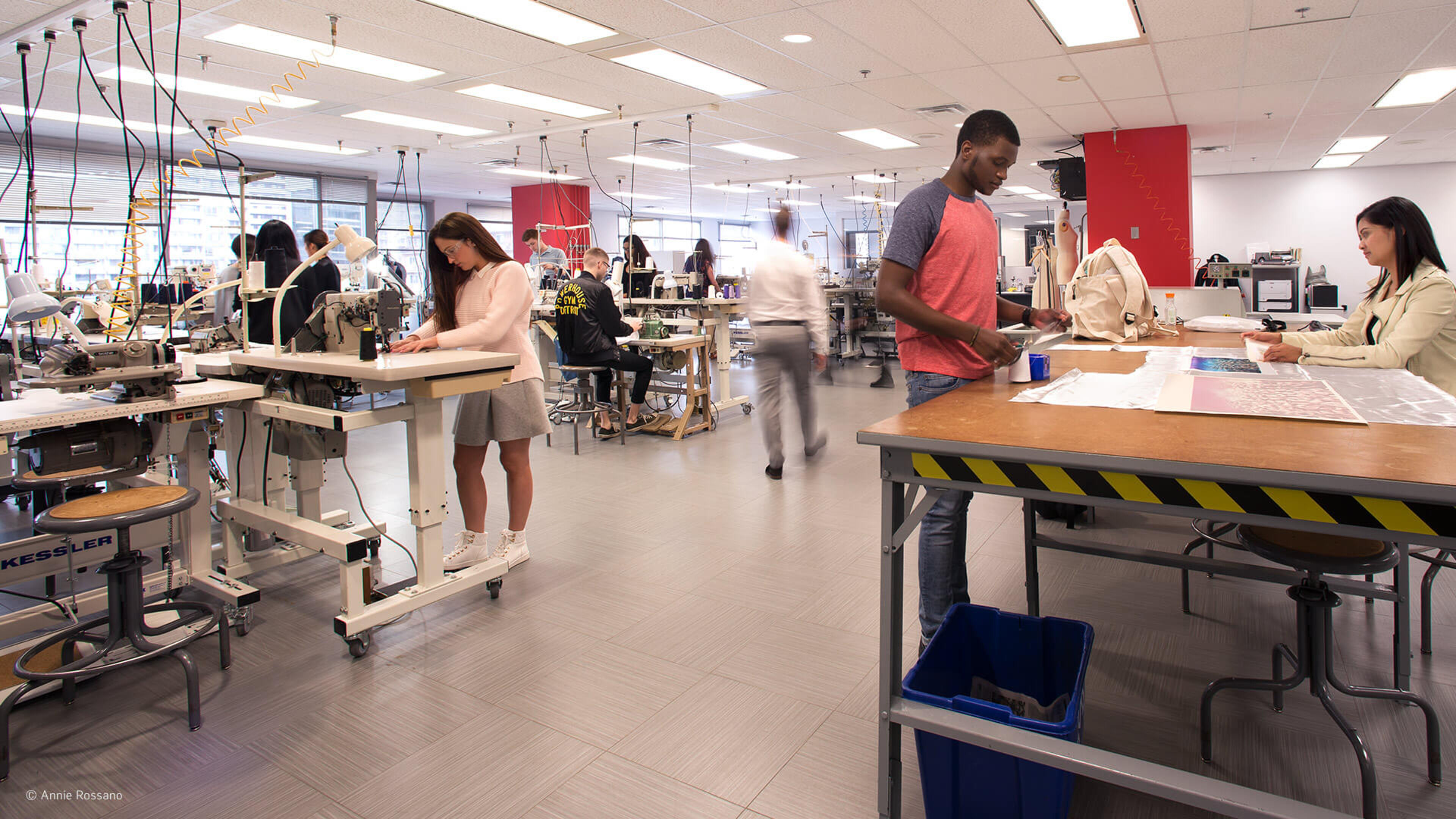 Fashion design students are actively working on projects in a well-equipped workshop with sewing machines and design stations.