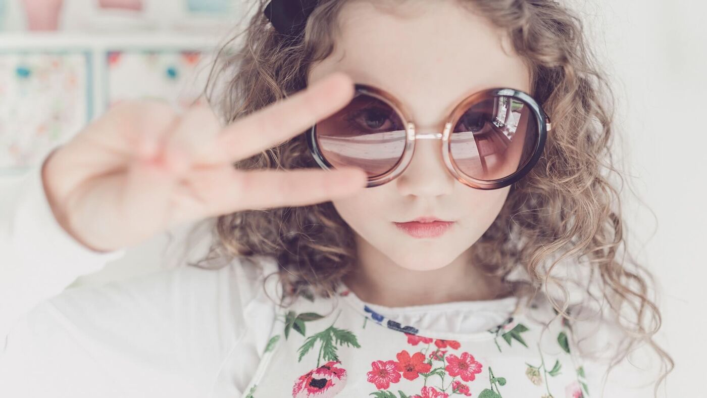 A young girl poses confidently, making a peace sign while wearing oversized sunglasses, portraying a playful and fashionable look.