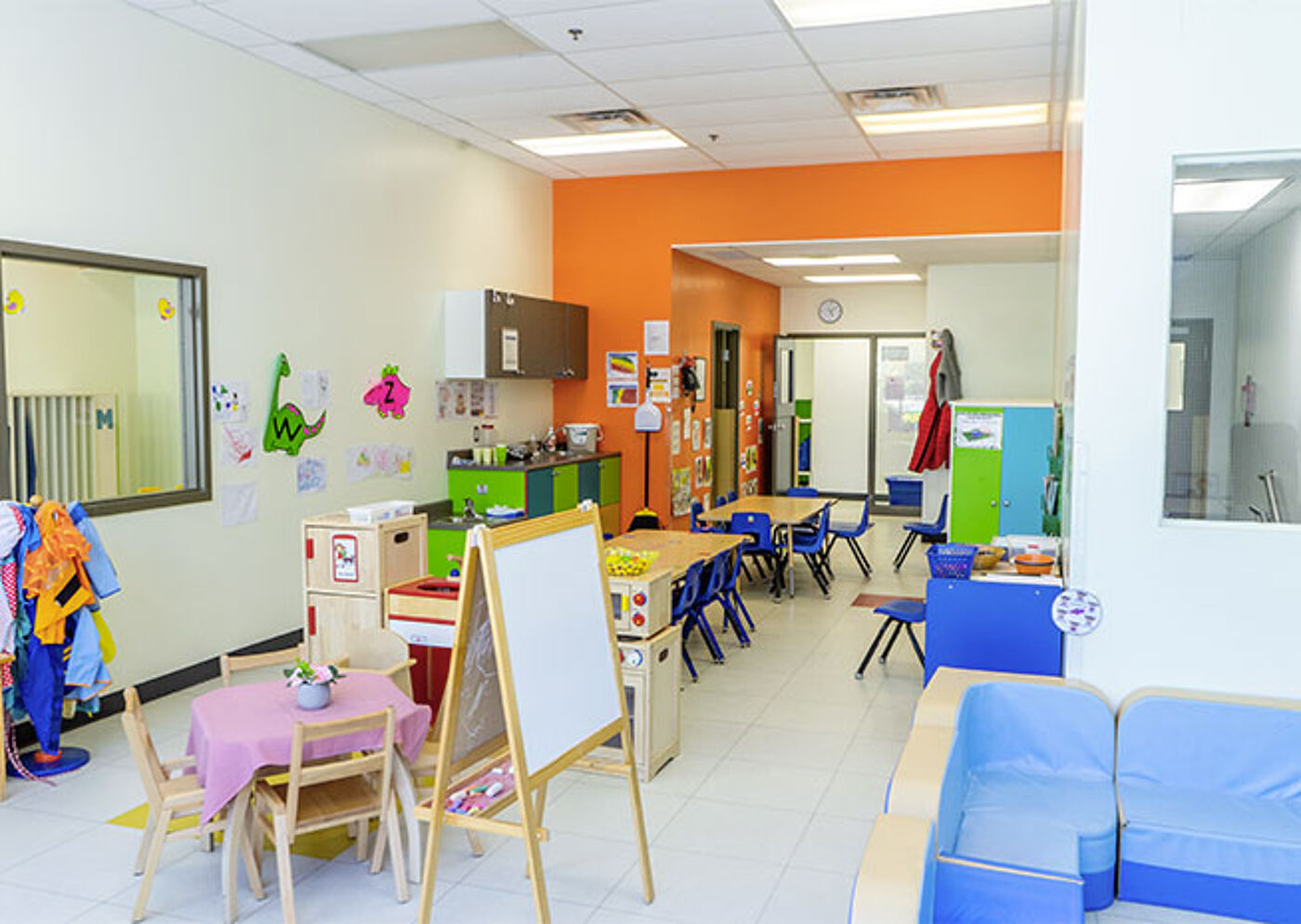 An orderly preschool classroom featuring a kitchen playset, a blue sofa, and blue chairs at tables, ready for learning activities.