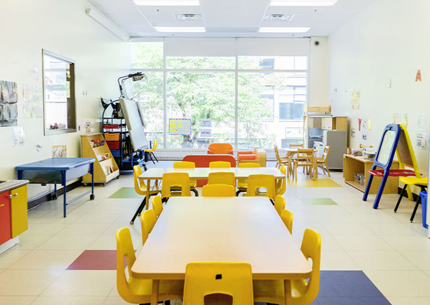 Large preschool classroom with yellow tables and chairs, and an array of educational materials, under natural light.