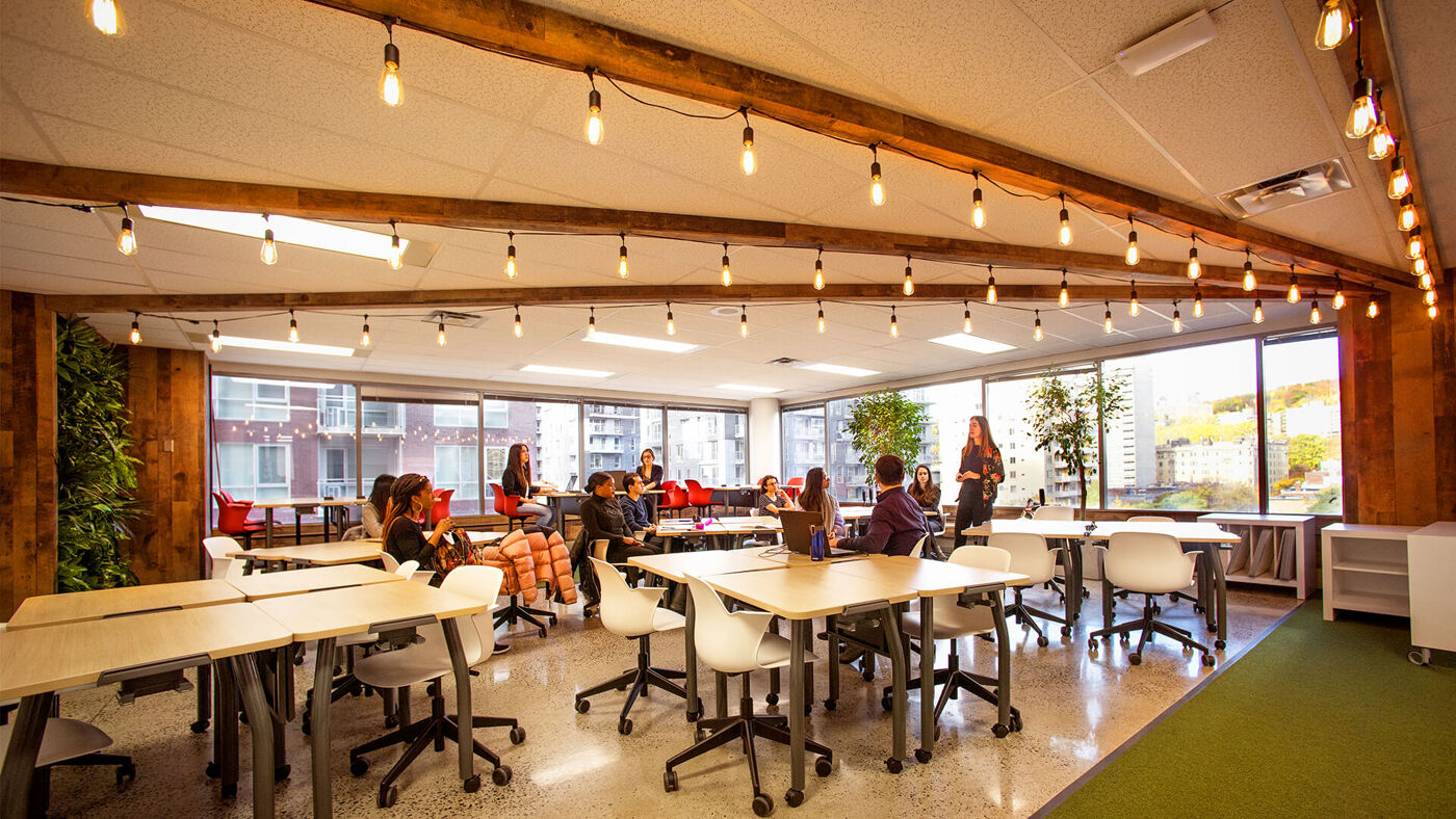 People engage in work and discussions in a spacious, well-lit co-working area with large windows and wooden accents.