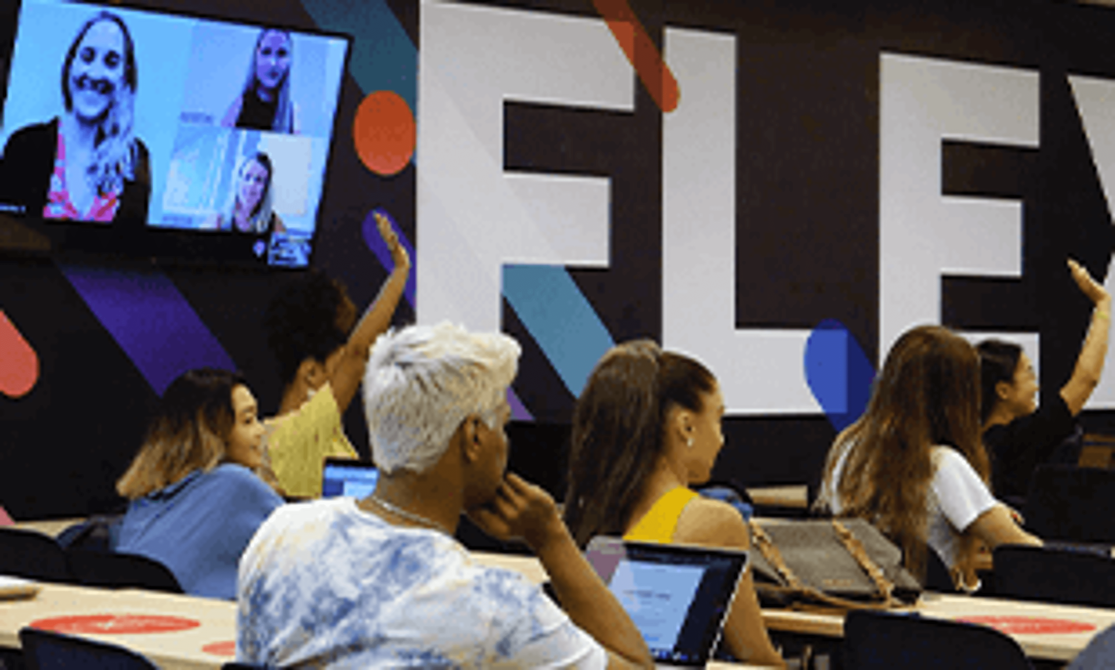 Students participate in an interactive session with a virtual instructor displayed on large screens in a modern classroom.