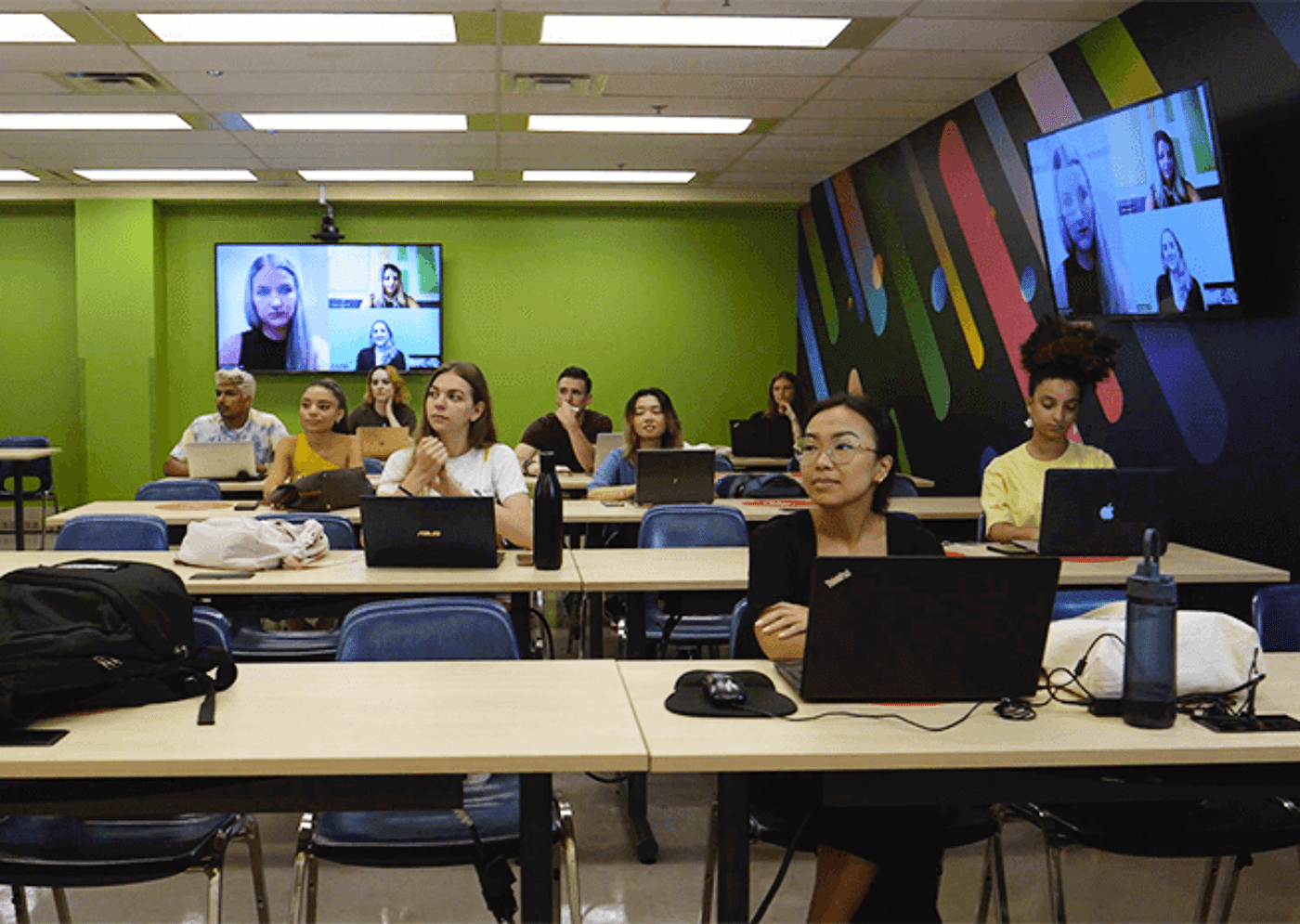 Students in a classroom with laptops, participating in a hybrid learning session with remote peers on a screen.