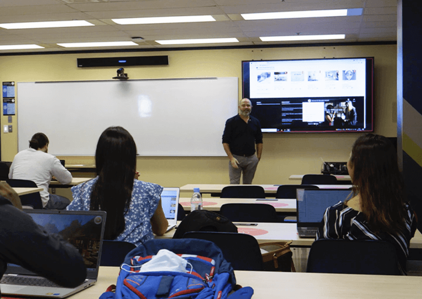 University students with laptops in a lecture hall, focusing on a professor near a presentation screen.