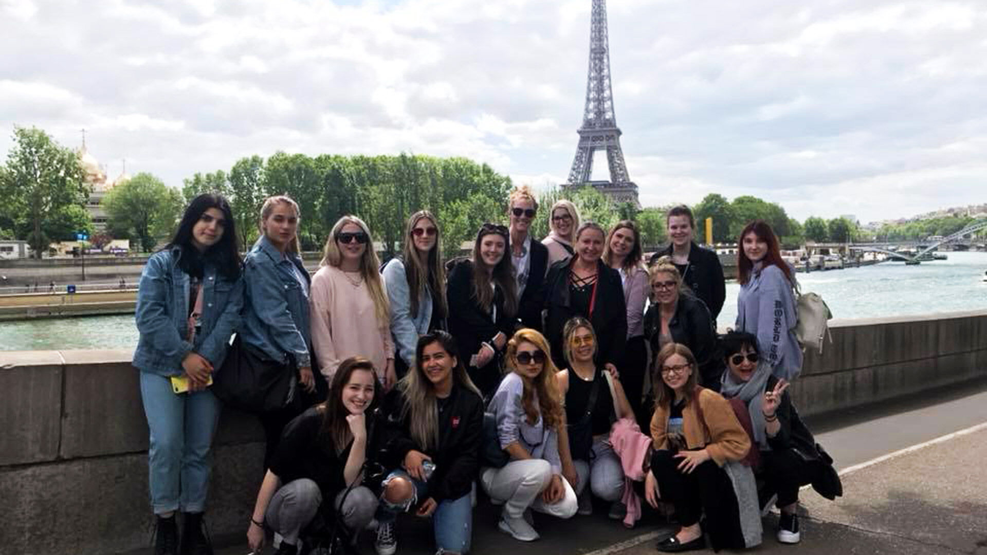 A group of tourists with the Eiffel Tower in the background, enjoying a sunny day by the Seine River in Paris.