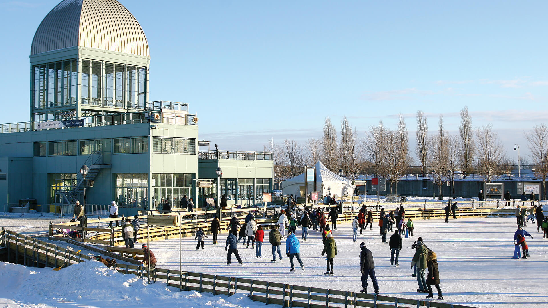 Des personnes de différents âges font du patin à glace sur une grande patinoire extérieure à côté d'un bâtiment moderne sous un ciel bleu dégagé.