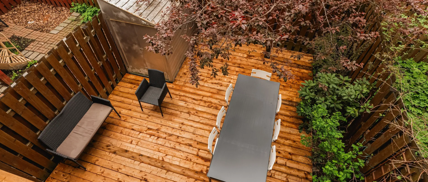 The image features a wooden deck with a dining table and chairs, surrounded by a garden with trees and a bird feeder.