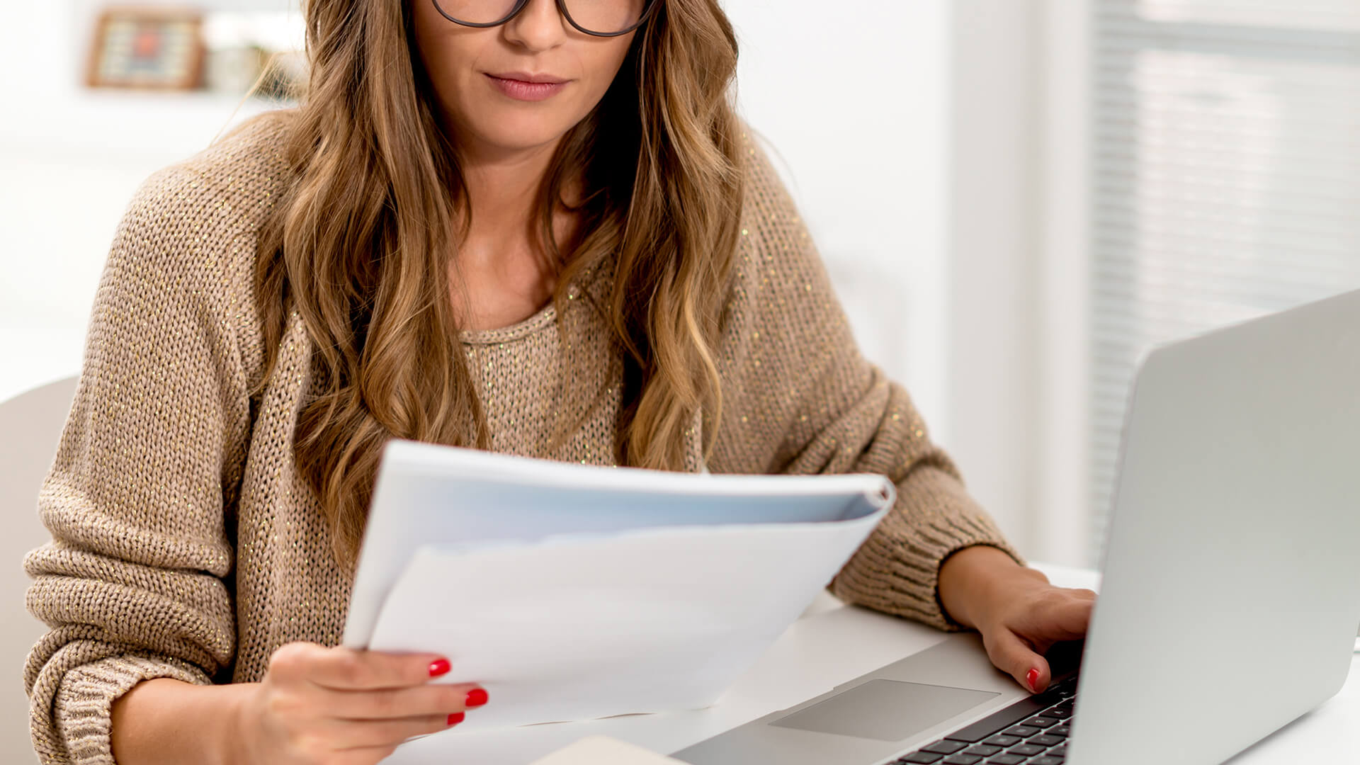 Une femme concentrée avec des lunettes examine des documents à côté de son ordinateur portable dans un bureau à domicile lumineux.