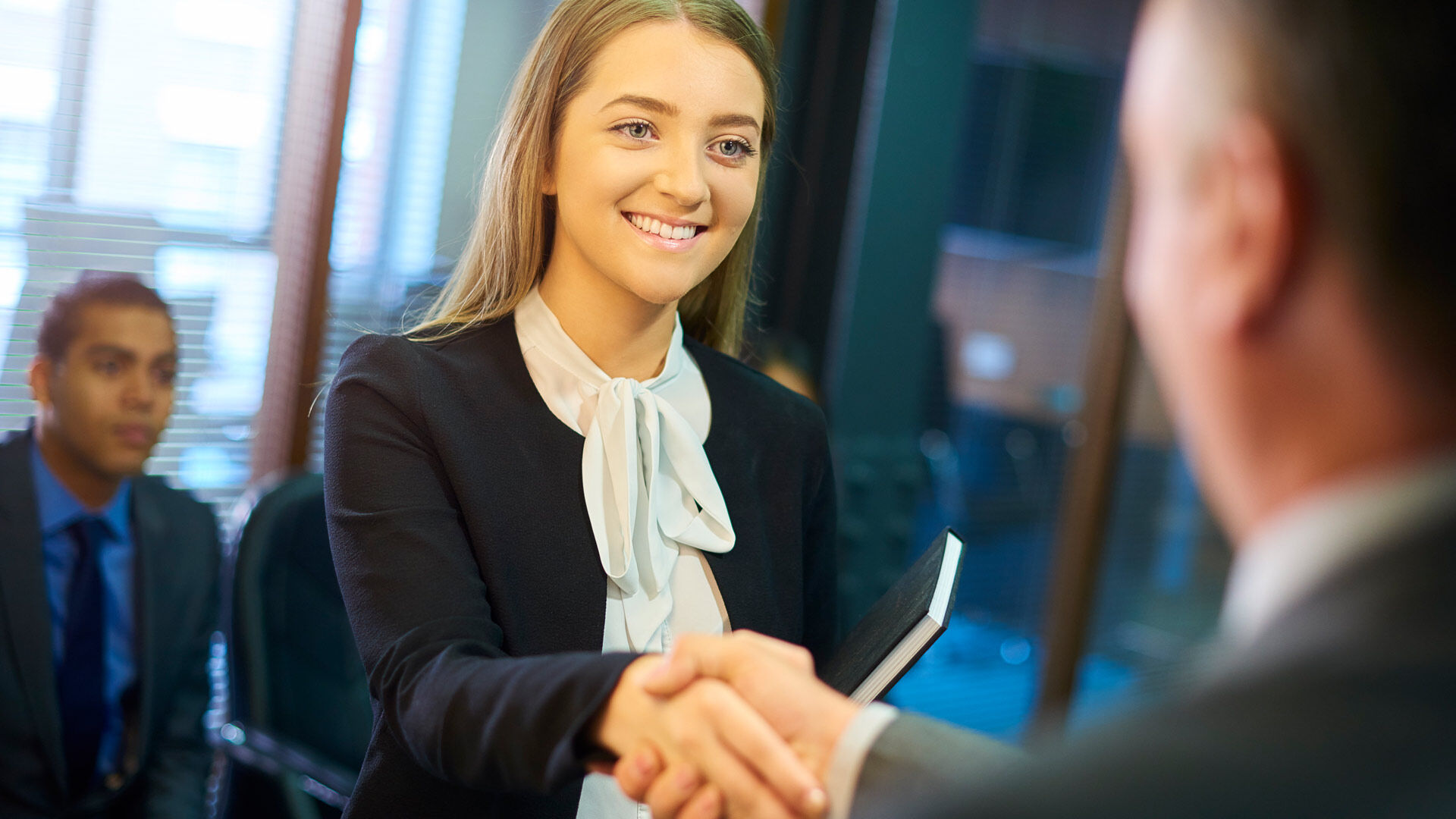 A woman in business attire shaking hands with a person, signaling a professional greeting or agreement in an office setting.