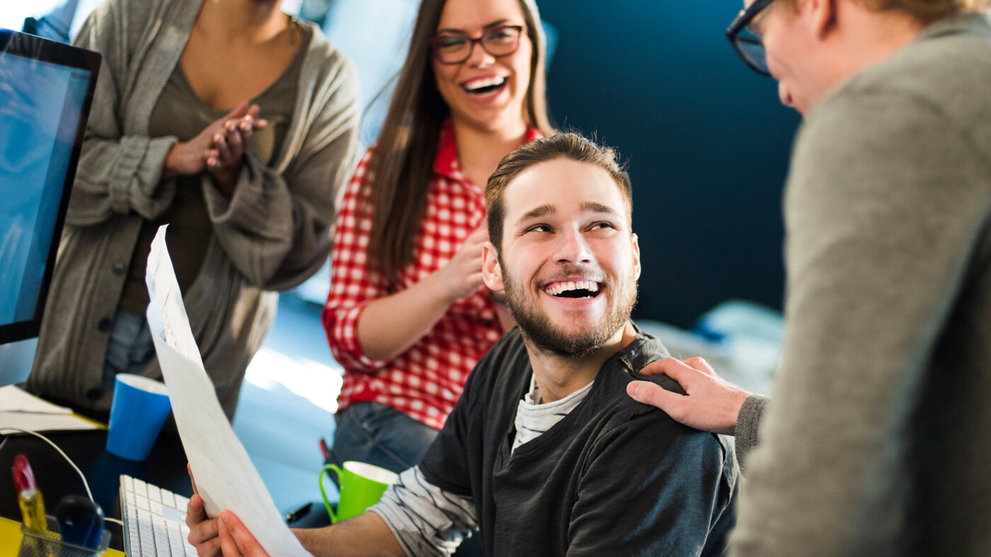 man in casual attire smiles brightly, surrounded by cheerful colleagues in an office setting, indicating a positive team dynamic.