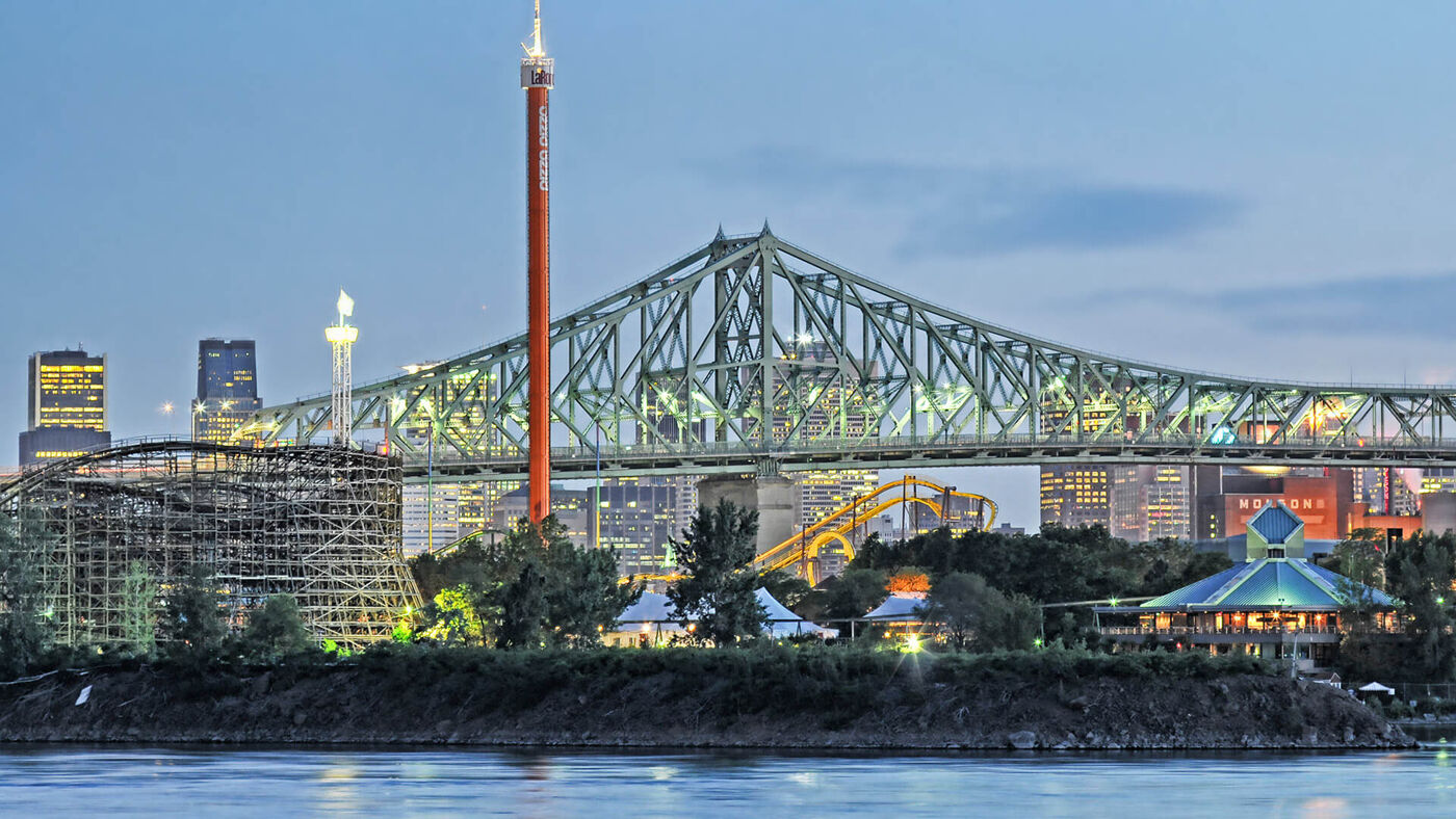 Twilight view of a steel truss bridge over a river, with an amusement park and urban skyline in the background.