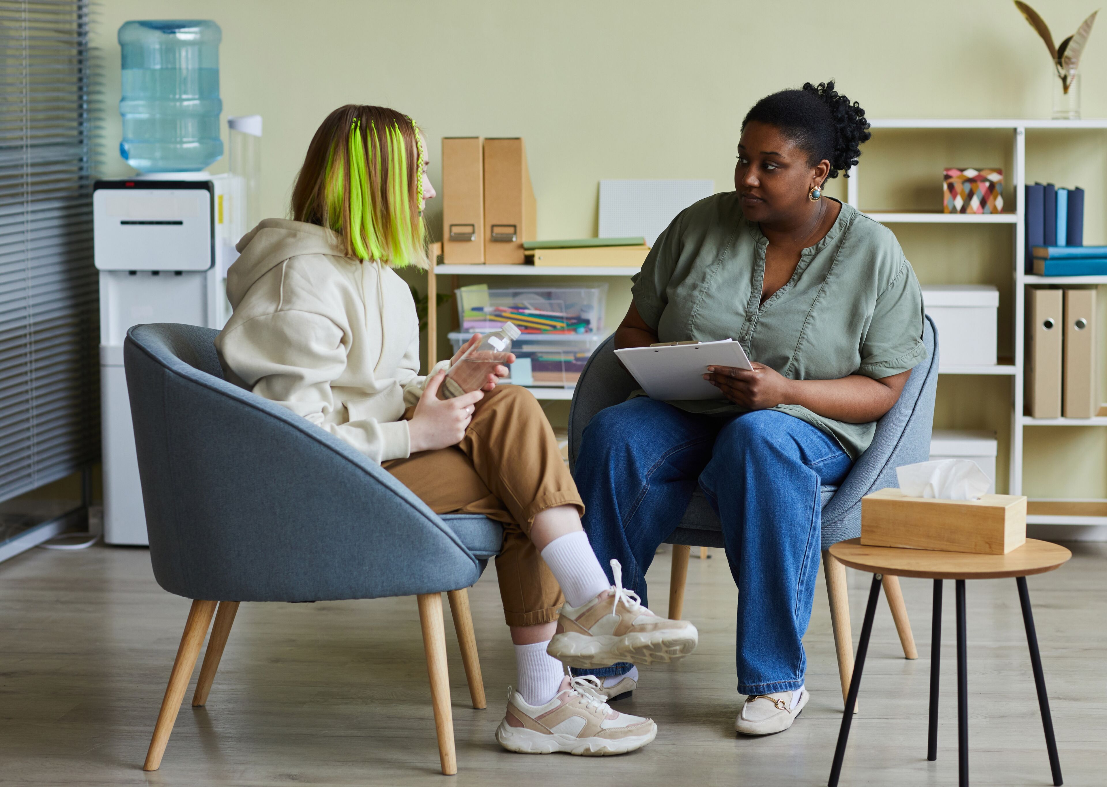 A patient with striking green hair speaks to a therapist, who is taking notes during a counseling session.