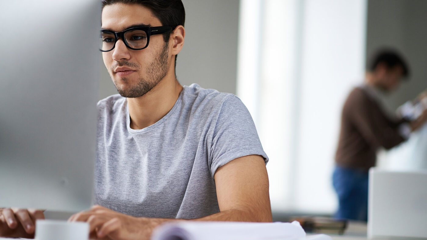 A young adult with glasses focused on paperwork at a modern workspace, with another individual blurred in the background