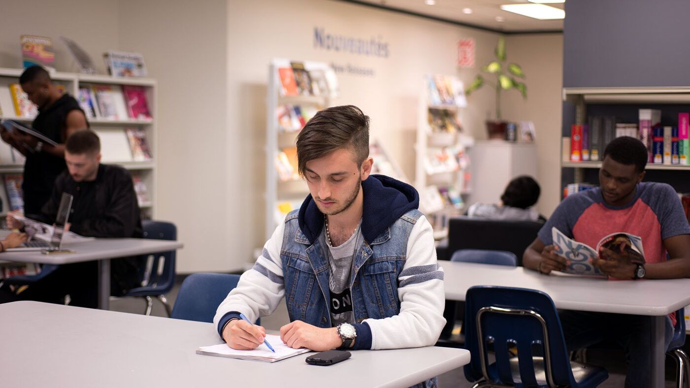 Three focused students engaged in reading and writing activities in a well-lit library setting.