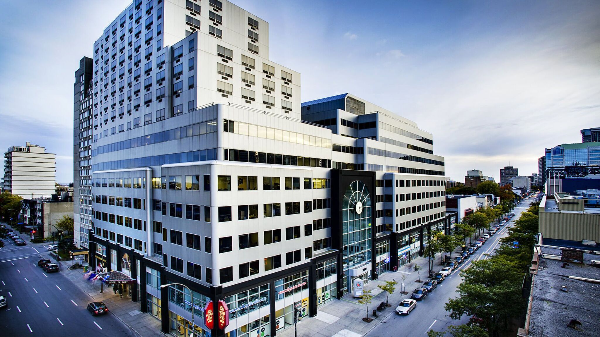 A cityscape at dusk featuring modern multi-story buildings with reflective windows, street-level shops, and sparse traffic on the adjacent streets.