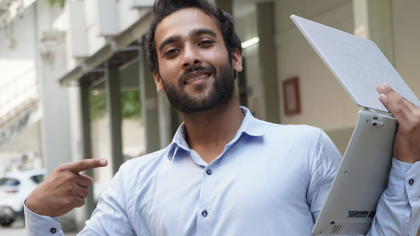 A cheerful young businessman in a light blue shirt points to himself, holding a grey laptop under his arm.