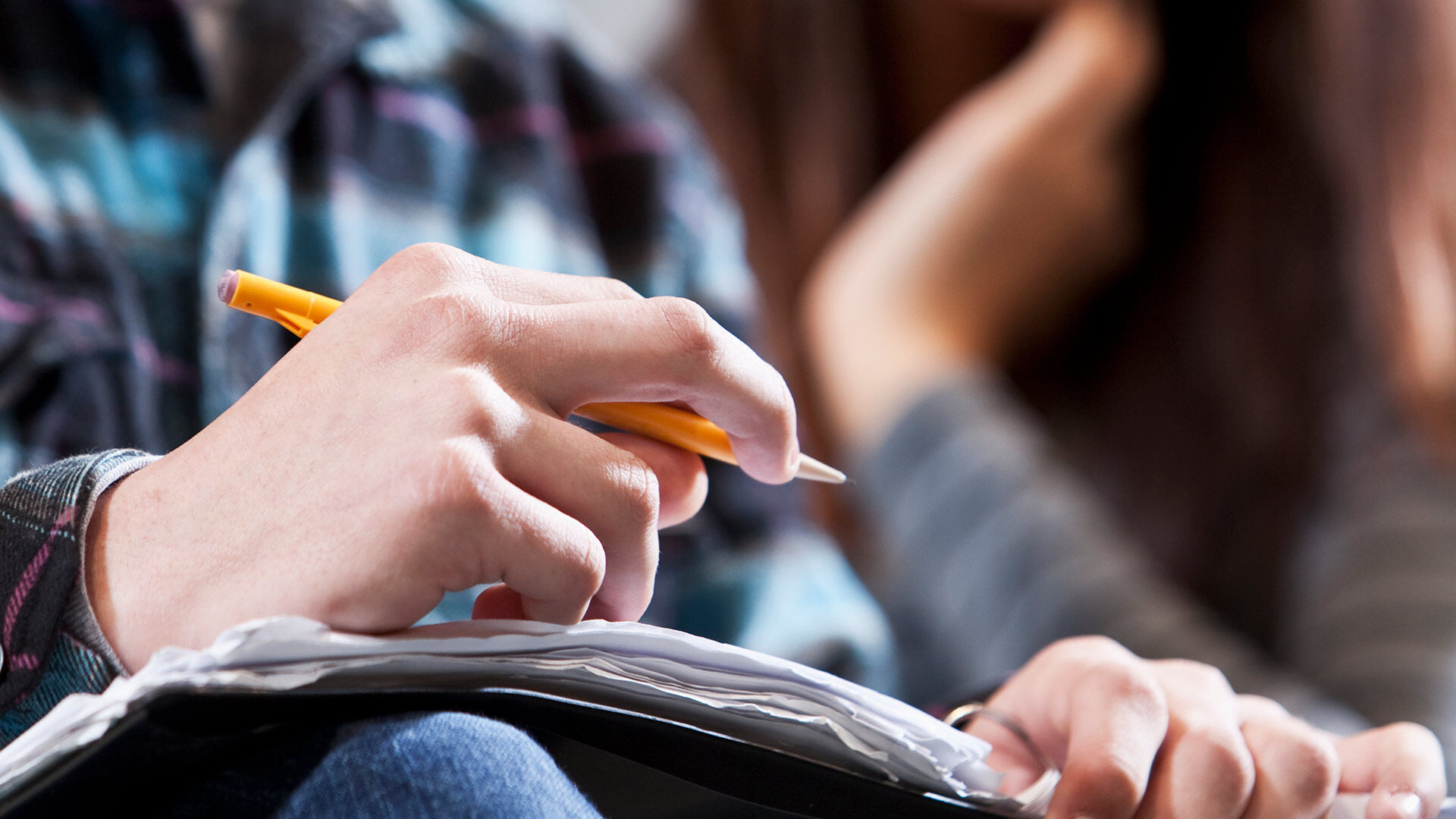 Gros plan sur la main d'un étudiant écrivant des notes avec un crayon jaune dans un cahier pendant un cours magistral.