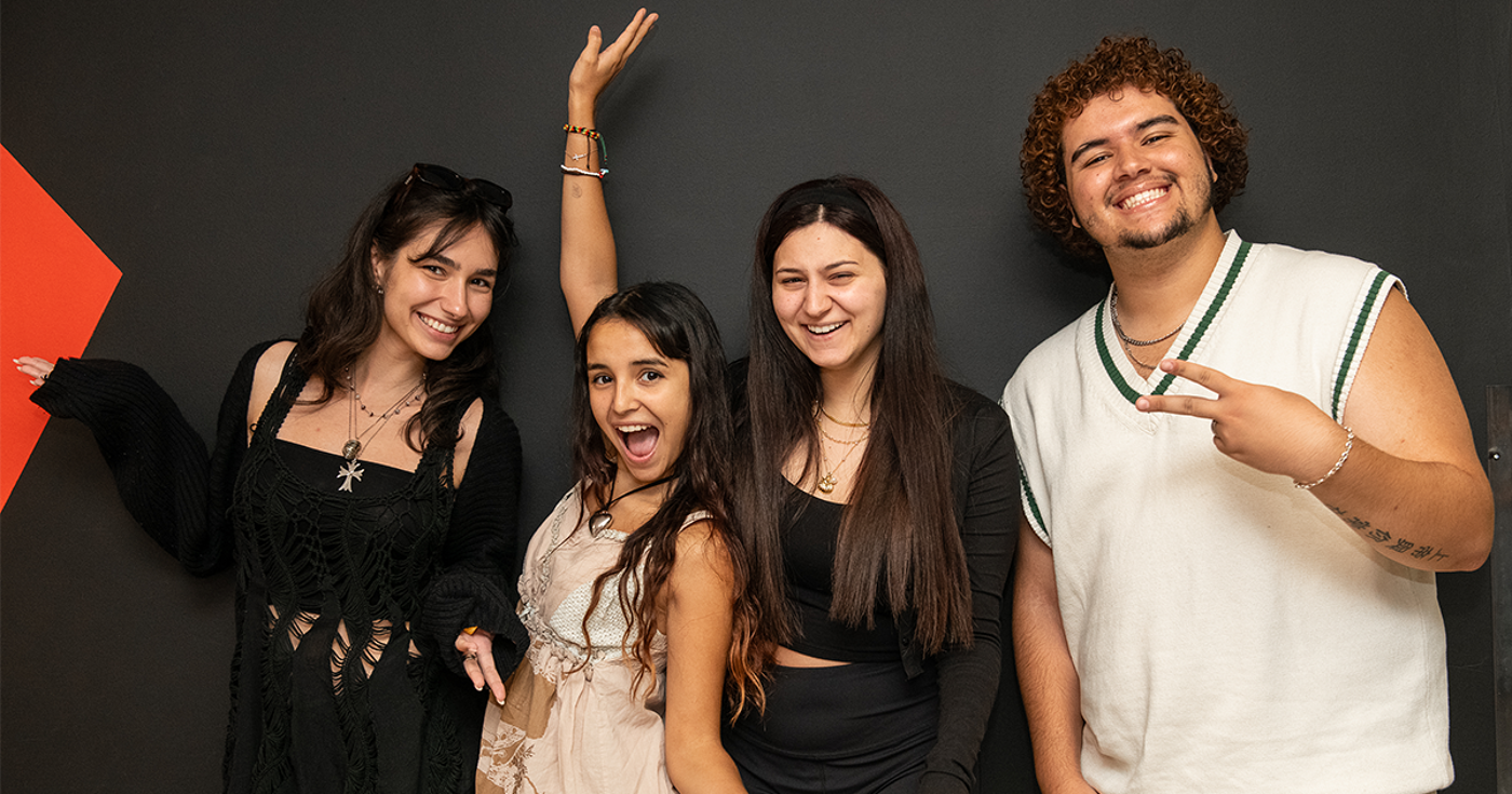 Four friends posing with cheerful expressions; two women raising their hands in a victory gesture, another smiling broadly, and a man making a peace sign.