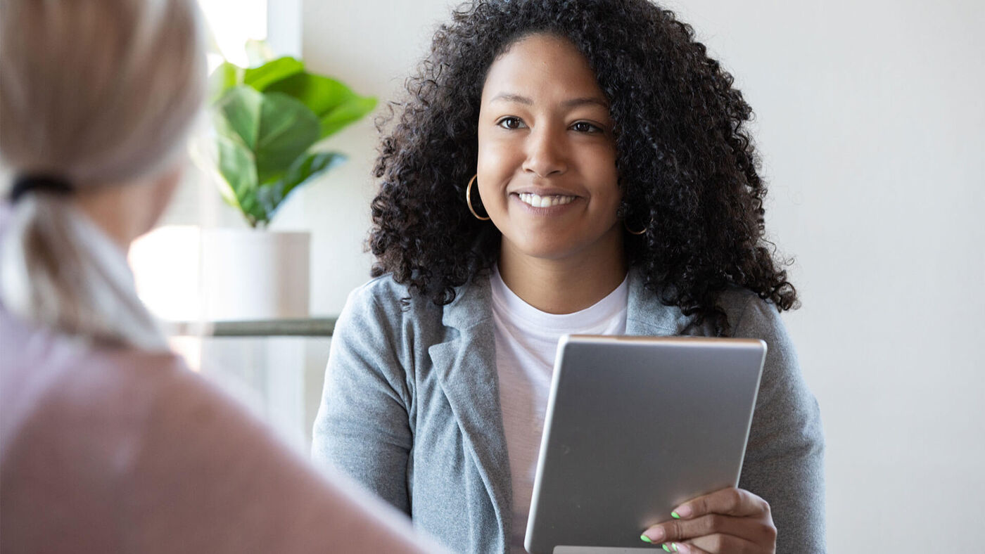 A young woman with curly hair smiles engagingly during a consultation, holding a digital tablet and actively listening, set against a bright, modern office backdrop.