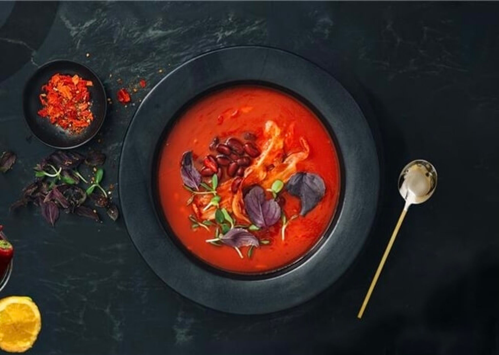Rich tomato soup garnished with herbs and beans, served in a black bowl on a dark surface.