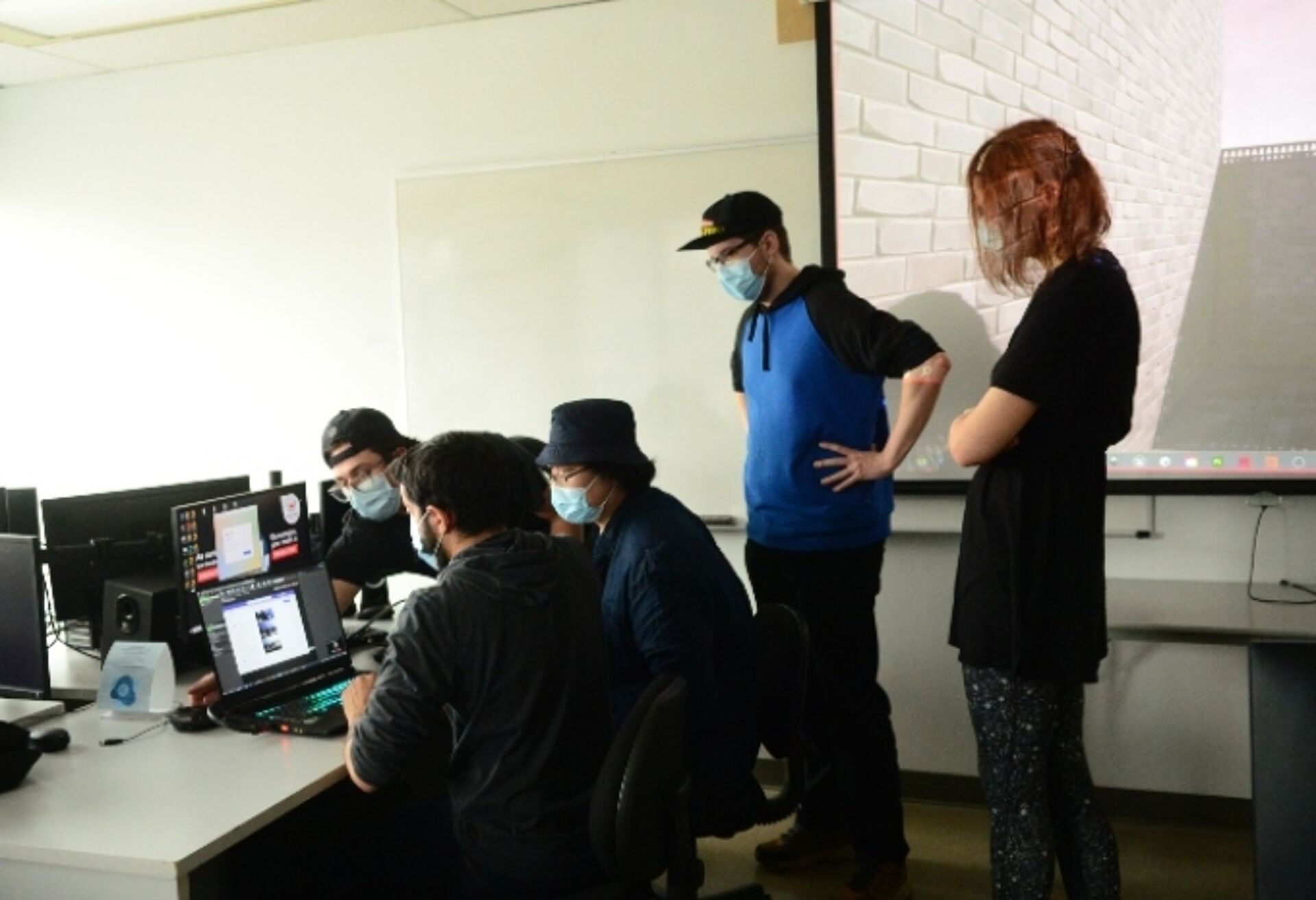 A team of tech professionals wearing masks collaborates at a workstation in an office environment.
