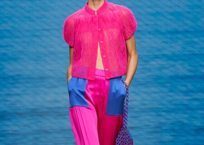 Un mannequin présente une déclaration de mode audacieuse avec une veste courte en fausse fourrure rose vif, un pantalon en satin bleu électrique et un sac assorti à maillons bleus.