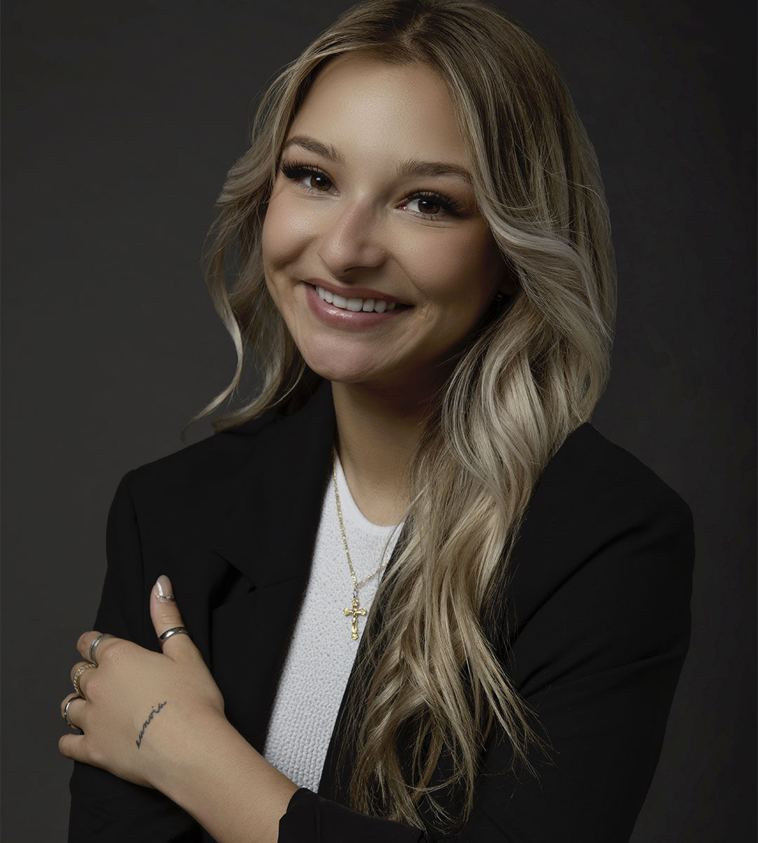 Une femme joyeuse aux cheveux blonds ondulés, portant un blazer noir sur un haut blanc, pose avec un sourire confiant.