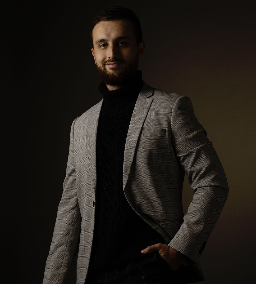 A poised young man stands with one hand in his pocket, wearing a sophisticated grey blazer over a black turtleneck, against a dark background.