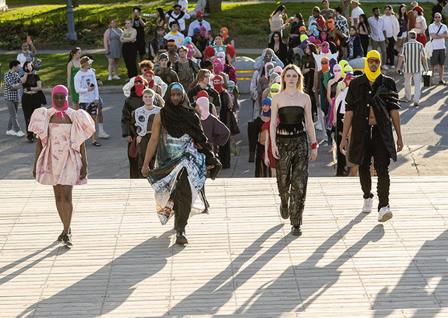 Models walk down a runway outdoors, showcasing diverse clothing styles in front of an audience.