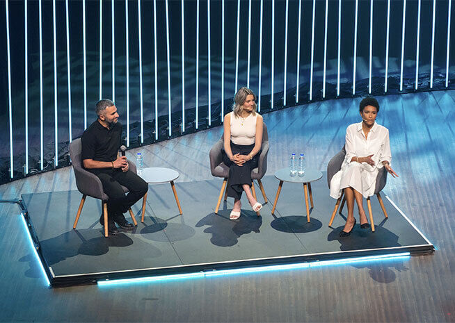 Trois professionnels assis sur scène lors d'une discussion de panel dans une conférence, avec un fond rayé moderne illuminé.
