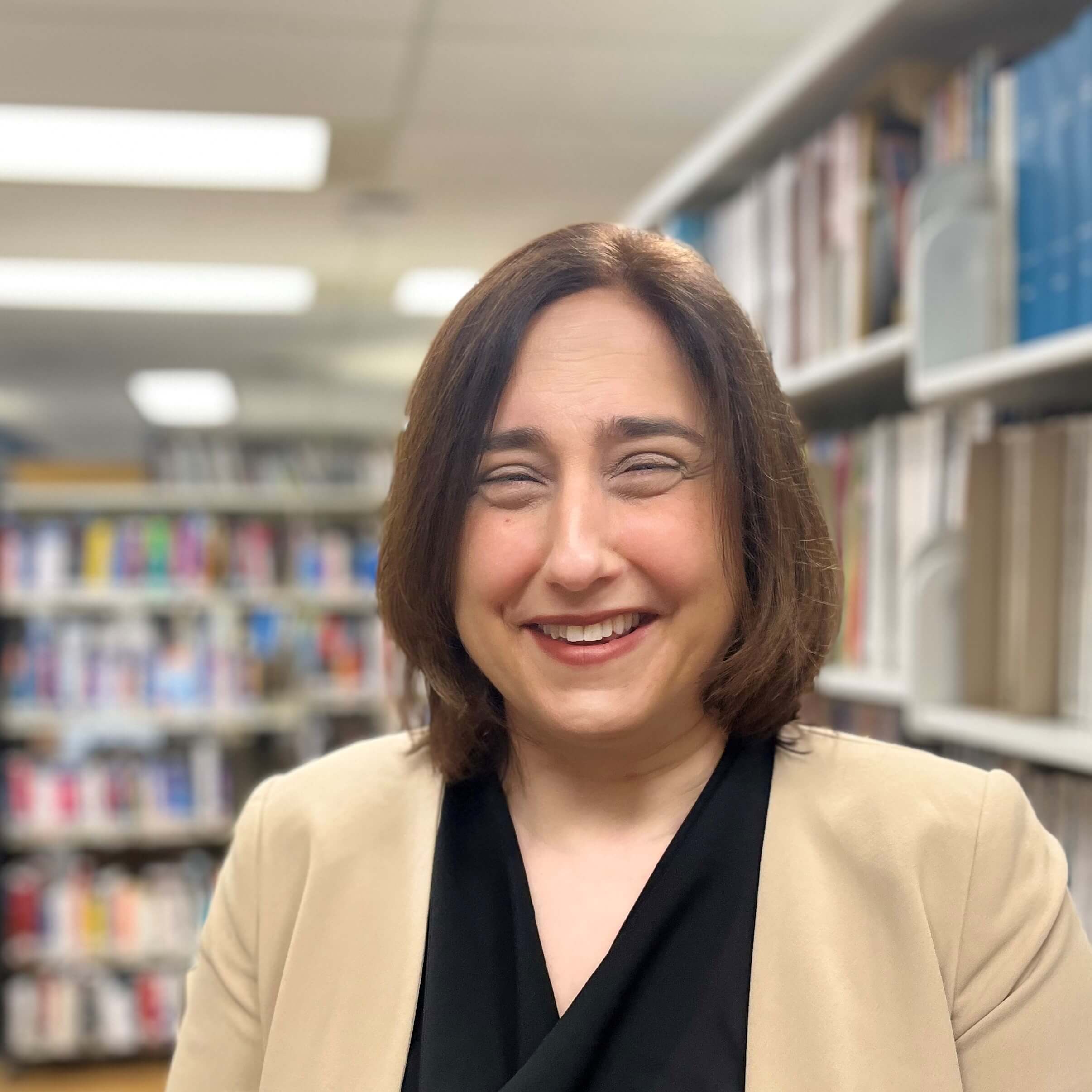 Femme satisfaite aux cheveux bruns mi-longs, souriant les yeux fermés, portant un blazer beige sur un haut noir, devant des étagères de livres.