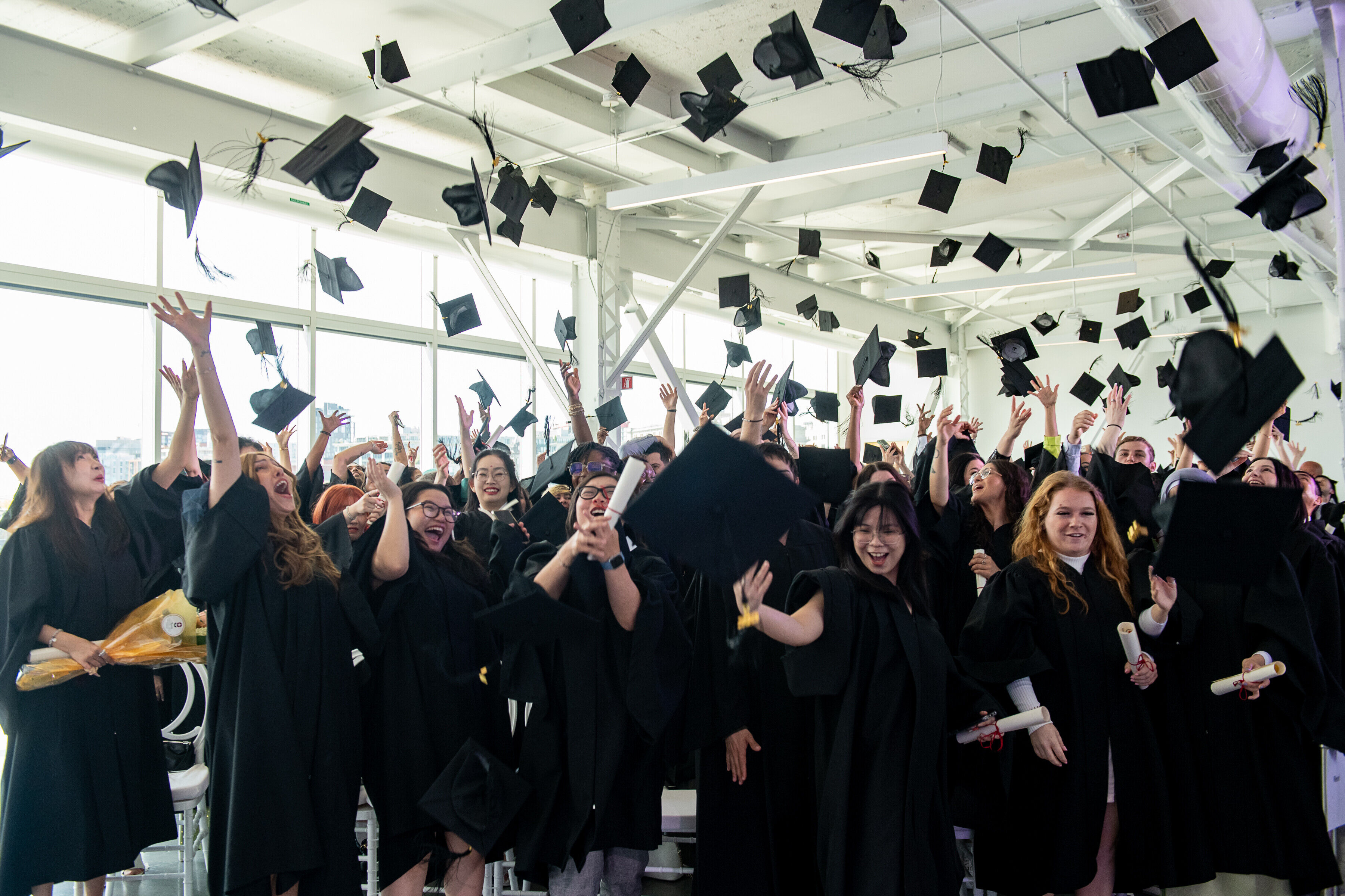 Un groupe de diplômés joyeux en toges et coiffes noires célèbrent en jetant leurs chapeaux en l'air dans une salle lumineuse.