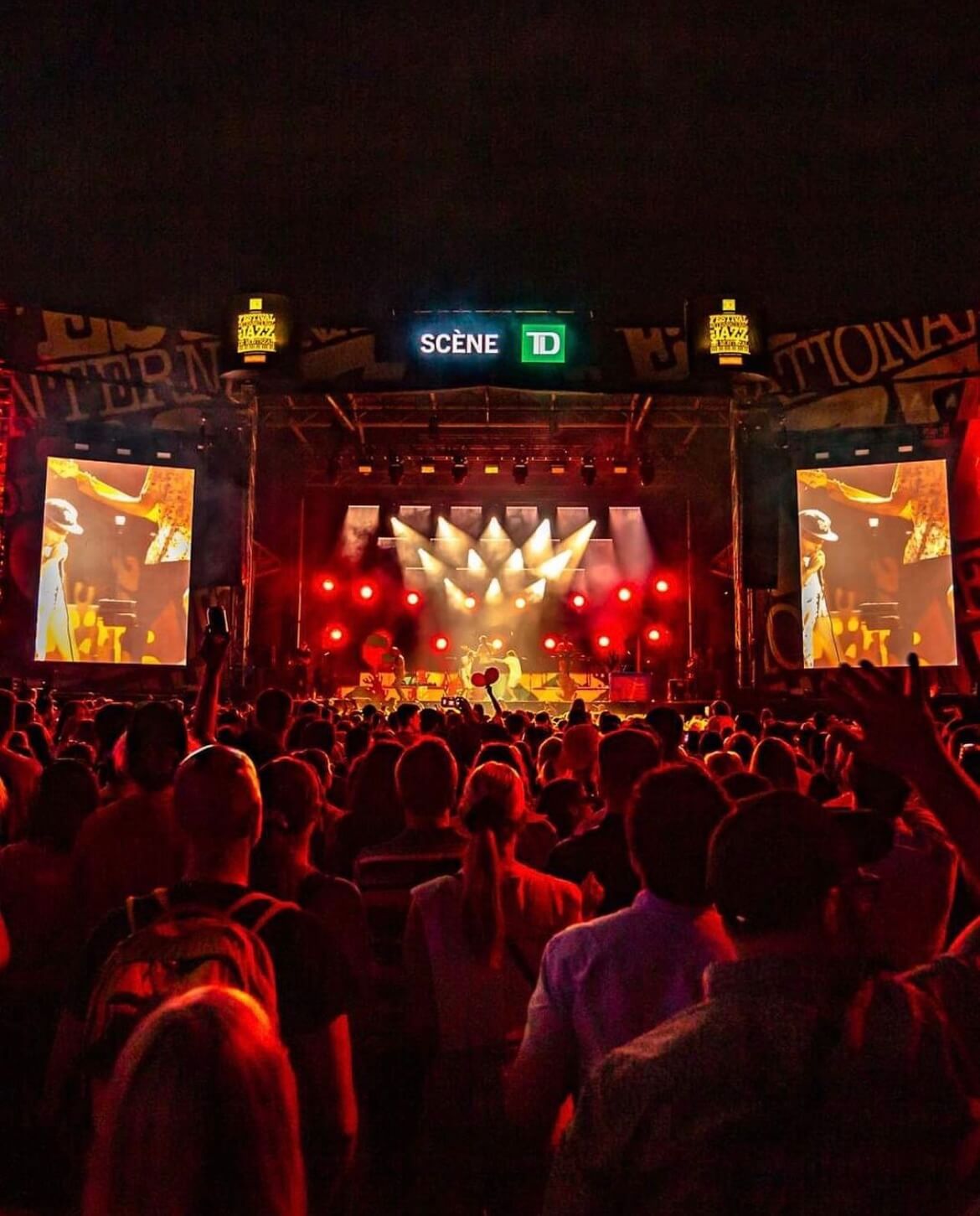 A lively crowd enjoying an outdoor concert, with vibrant stage lights illuminating the performers at a festival sponsored by TD.