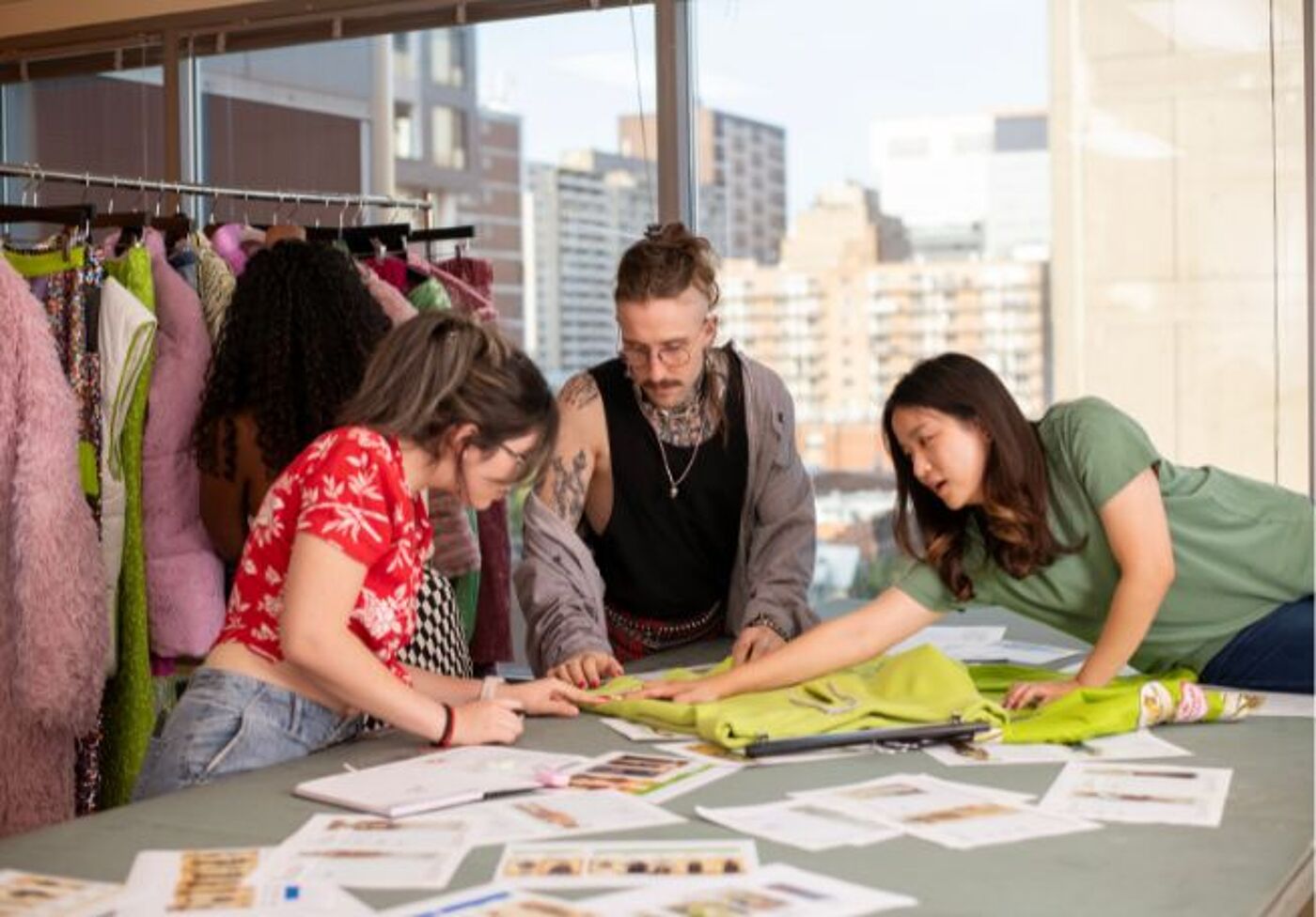 A diverse group of fashion designers collaborates on a project, discussing fabric choices and designs on a busy studio table, surrounded by vibrant clothing samples.