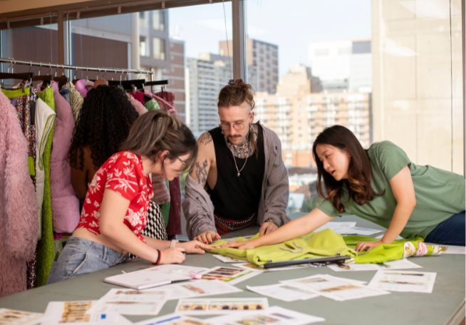 Un groupe diversifié de créateurs de mode collabore sur un projet, discutant des choix de tissus et des designs sur une table de studio animée, entourée d'échantillons de vêtements vibrants.