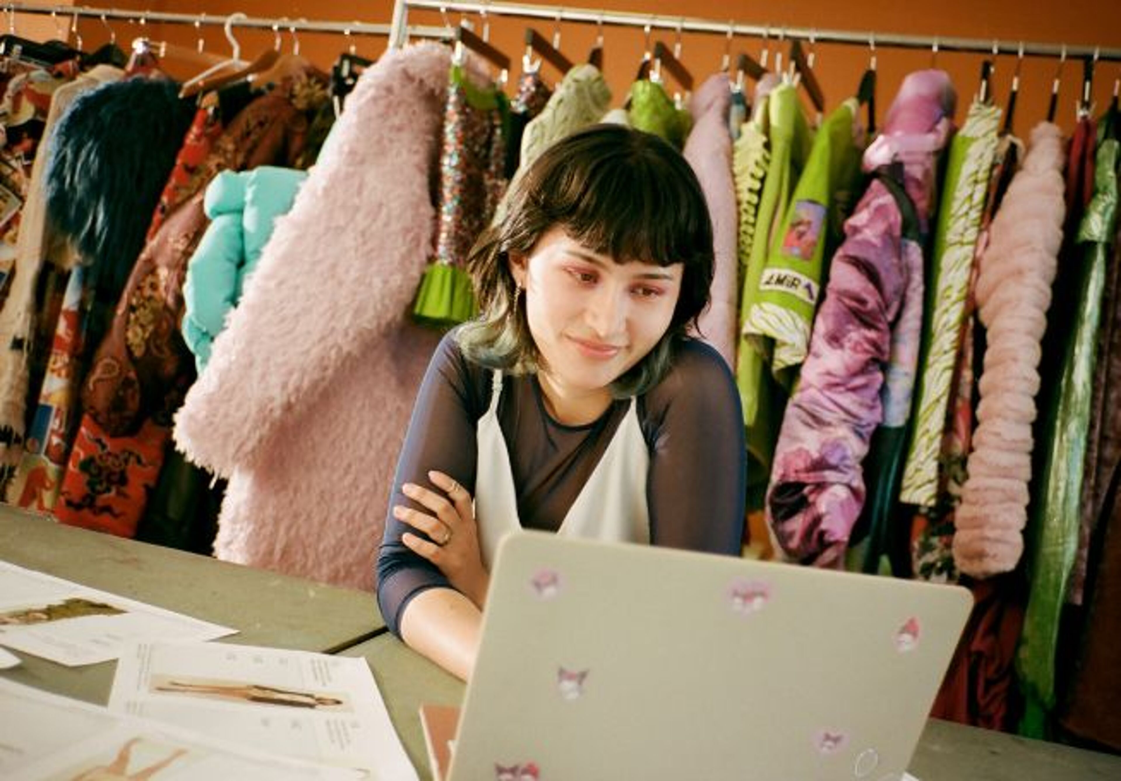 A young fashion designer focused on her laptop in a vibrant studio, surrounded by a colorful array of textured garments hanging in the background.