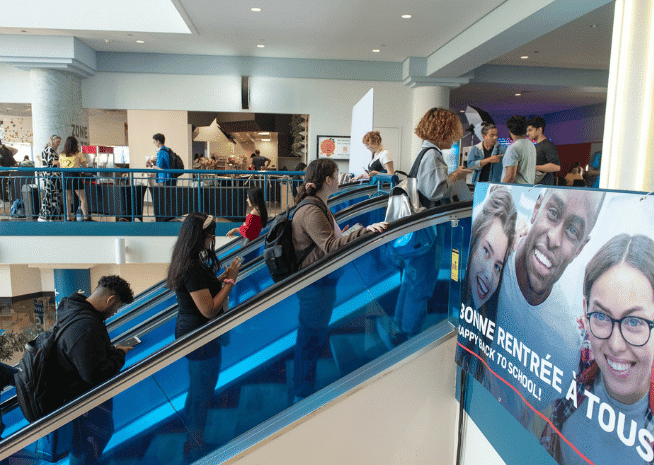 Students moving up and down an escalator in a busy college building, with banners promoting a back-to-school campaign, reflecting the lively atmosphere of academic life.