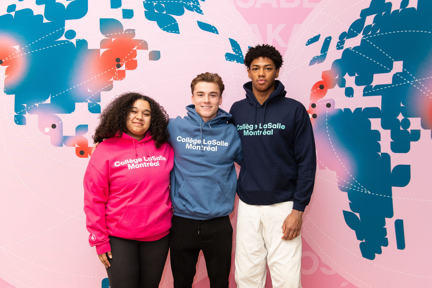In the picture, there are three people, two men and a woman, posing together in front of a map. They are wearing hoodies and smiling for the camera.
