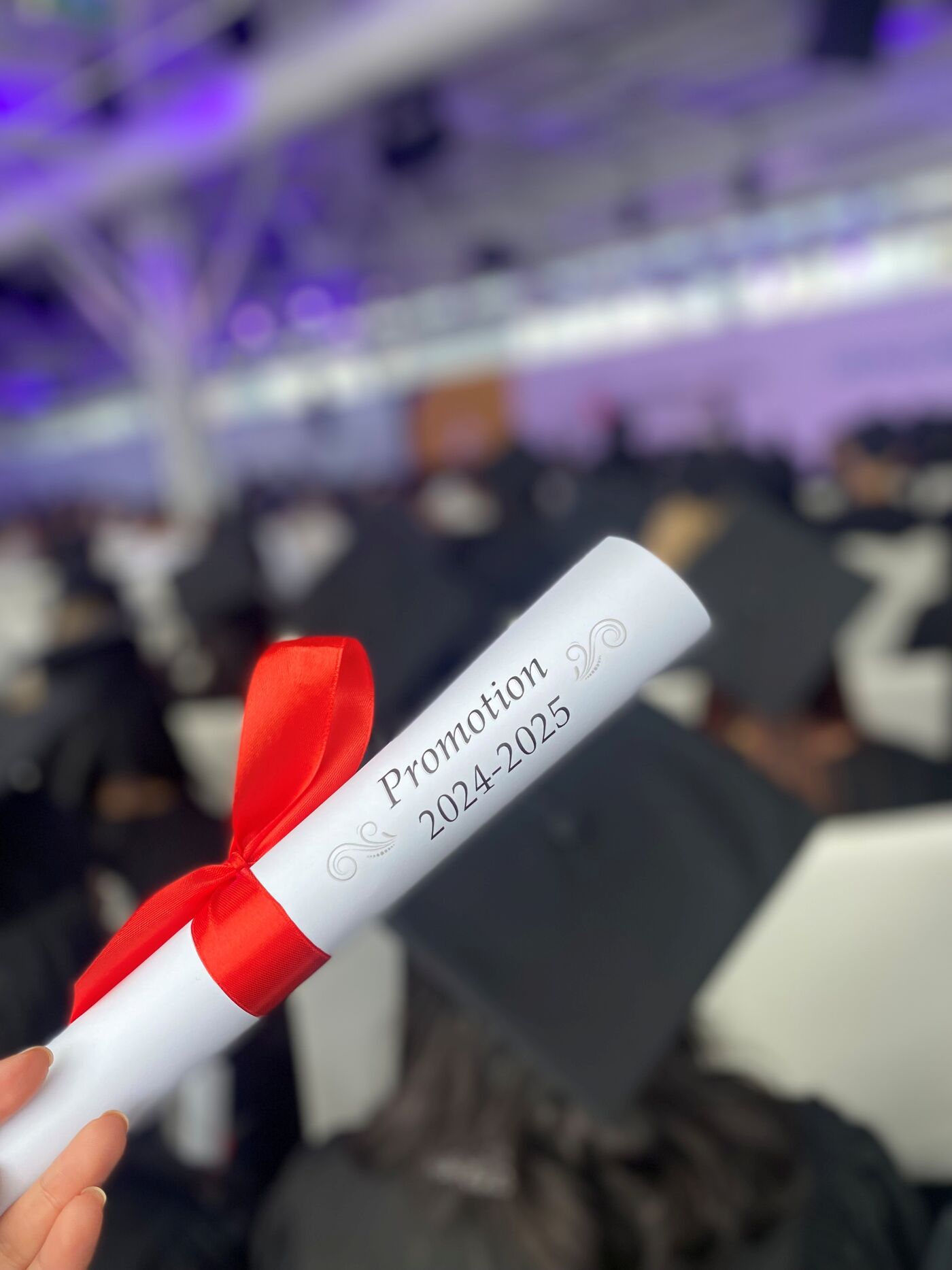 Close-up view of a hand holding a white scroll tied with a red ribbon, with "Promotion 2024-2025" printed on it, set against a blurred background of a graduation ceremony.