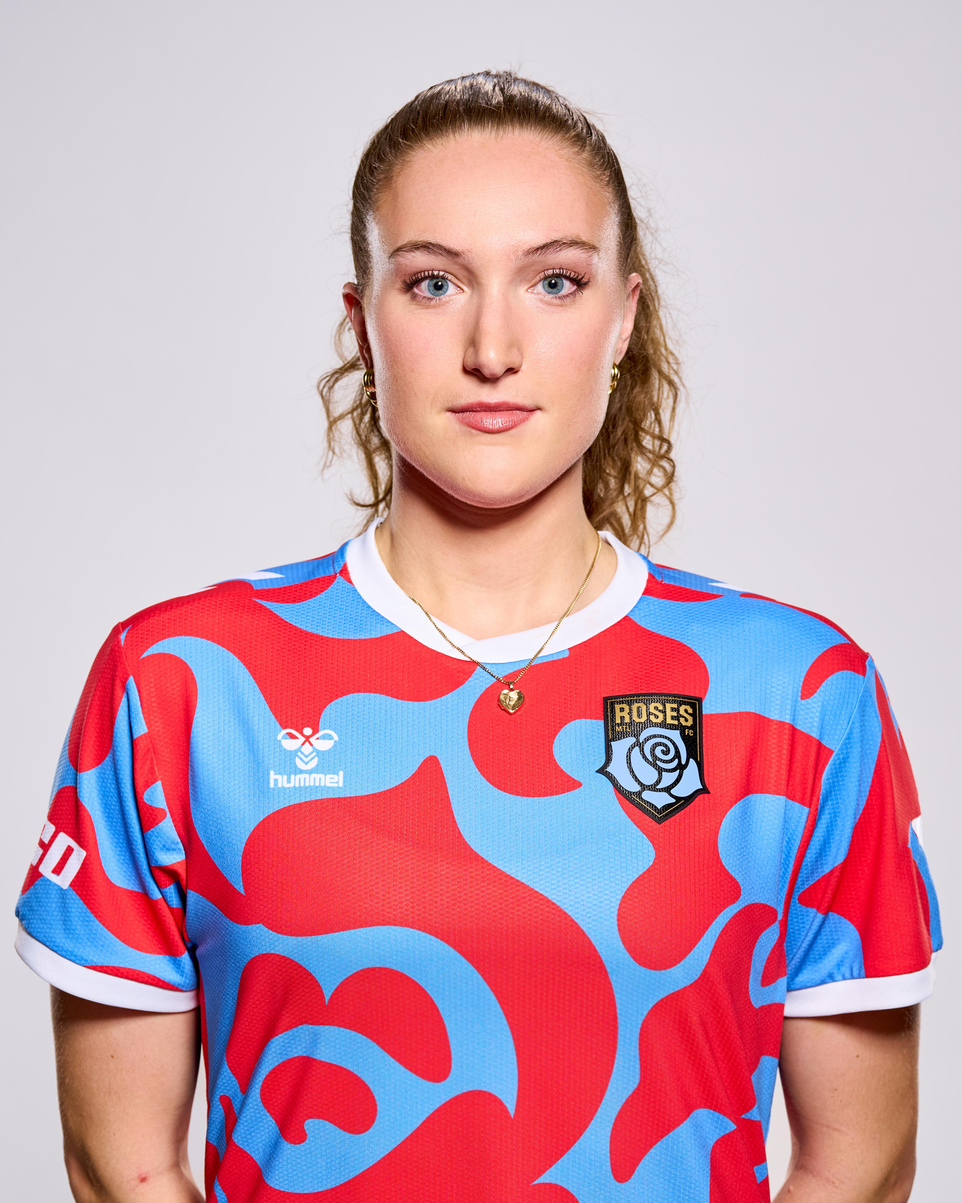 This is a photograph of a woman with her hair pulled back in a ponytail, wearing a short-sleeved jersey with a red and blue design. She is wearing a delicate necklace with a small pendant.